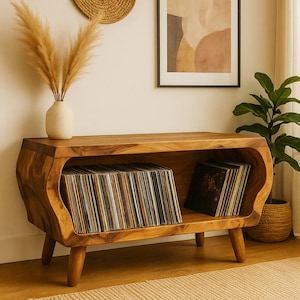 May include: A wooden record player stand with a curved design. The stand is made of light brown wood and has a shelf for storing vinyl records. A vase with dried flowers sits on top of the stand.