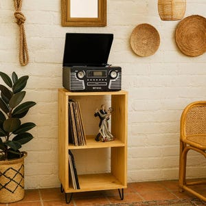 Op de afbeelding: Een zwart-zilveren platenspeler in retrostijl staat op een licht houten plank. De plank bevat vinylplaten en een beeldje van een dansend stel. De unit heeft zwarte haarspeldpoten en staat tegen een witte bakstenen muur.