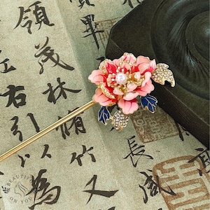 May include: A decorative hair stick featuring a pink and red flower with a pearl center, blue leaves, and gold accents. The flower is adorned with rhinestones. The stick is gold-colored. The background is a green fabric with Asian calligraphy.