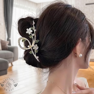 Può includere: Una molletta per capelli decorativa dorata con un design floreale. Presenta fiori bianchi, foglie verdi e perle finte. La molletta tiene i capelli castano scuro raccolti in uno chignon.