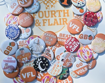 Könnte beinhalten: Eine Sammlung runder Buttons in verschiedenen Farben mit Text und Grafiken. Die Buttons zeigen Sätze wie "VFL", "Go Vols" und "Karma is a Vol". Einige Buttons haben Bilder eines Tigers und eines Footballspielers. Der Hintergrund lautet "FOURTH & FLAIR".