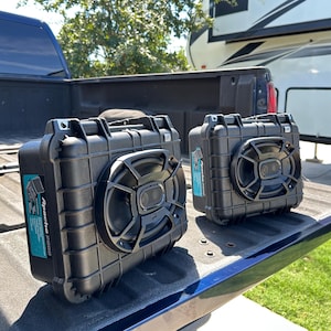 May include: Two black, rectangular speakers with a circular speaker grill are displayed on a blue surface. The speakers have the text "Apache 2000" on a blue label. A recreational vehicle is visible in the background.