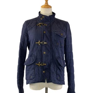 May include: A navy blue quilted jacket with a stand-up collar and brass-colored toggle closures. The jacket features two front pockets with flaps and quilted shoulder patches. The jacket is displayed on a mannequin.