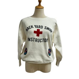 May include: Cream-colored sweatshirt with the text "BACK YARD SWIM INSTRUCTOR" above a red cross symbol. The sweatshirt has two small patches of a flip-flop design on the front.