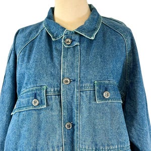 May include: A medium-wash denim jacket with a classic collar and button-up front. The jacket features two chest pockets with button closures and a tailored fit. The stitching is a contrasting light green color, and the buttons are silver.