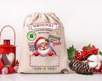 May include: A beige Christmas gift bag with a drawstring closure. The bag features a Santa Claus graphic and text that reads "North Pole Overnight Service." Decorative elements include a red lantern, berries, and a pinecone.