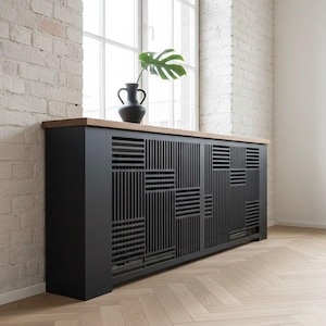 Könnte beinhalten: Ein schwarzer Heizkörperverkleidung mit geometrischem Design, gekrönt von einer hellen Holzoberfläche. Eine schwarze Vase mit einem grünen Blatt steht oben. Die Verkleidung befindet sich vor einem Fenster und einer weißen Backsteinmauer.