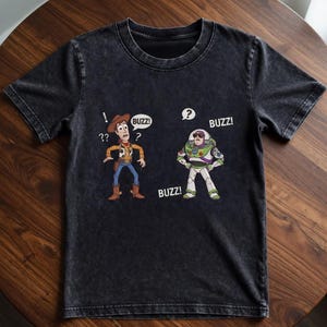 May include: Dark gray t-shirt featuring cartoon illustrations of Woody and Buzz Lightyear from Toy Story. Woody has a question mark above his head, and Buzz has a speech bubble with a question mark. The word "BUZZ!" appears in speech bubbles.