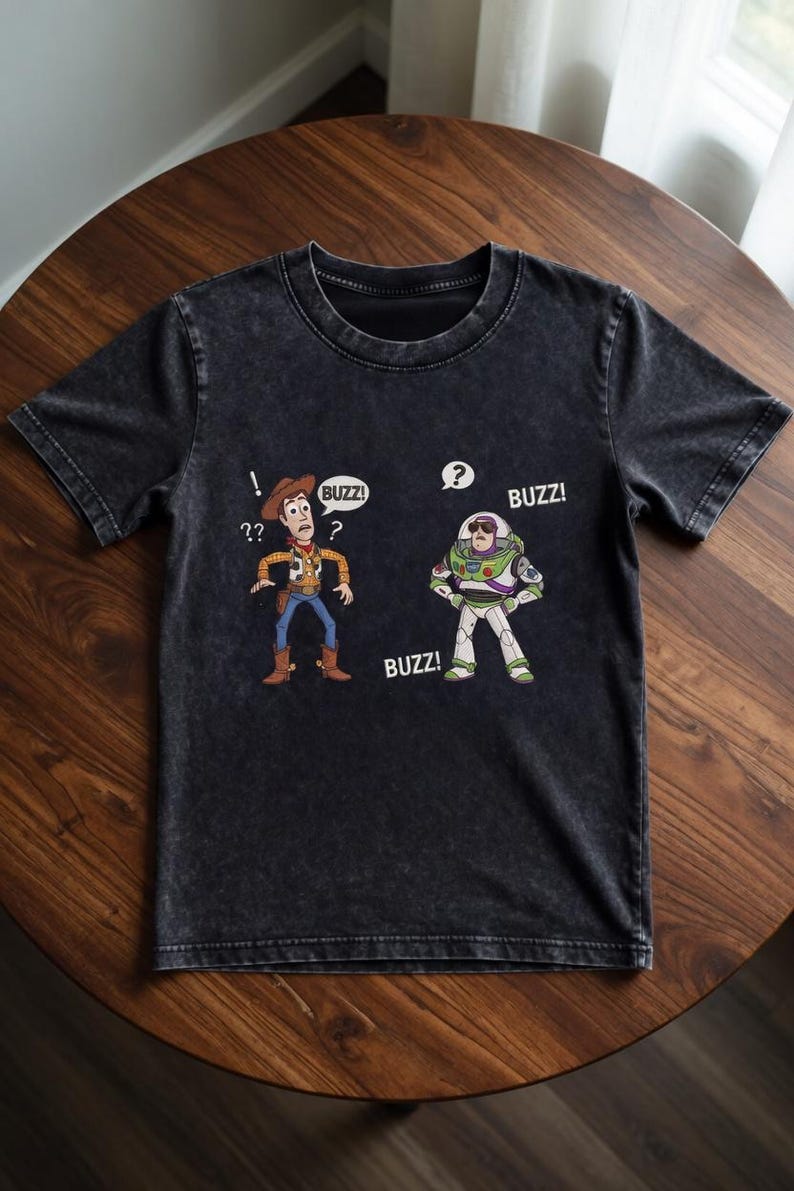 May include: Dark gray t-shirt featuring cartoon illustrations of Woody and Buzz Lightyear from Toy Story. Woody has a question mark above his head, and Buzz has a speech bubble with a question mark. The word "BUZZ!" appears in speech bubbles.