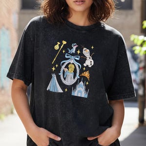 May include: Dark grey t-shirt with embroidered images of a princess, a snowman, a castle, and other elements from a popular animated film. The design includes a gold scepter, a blue dress, and a blue bow.