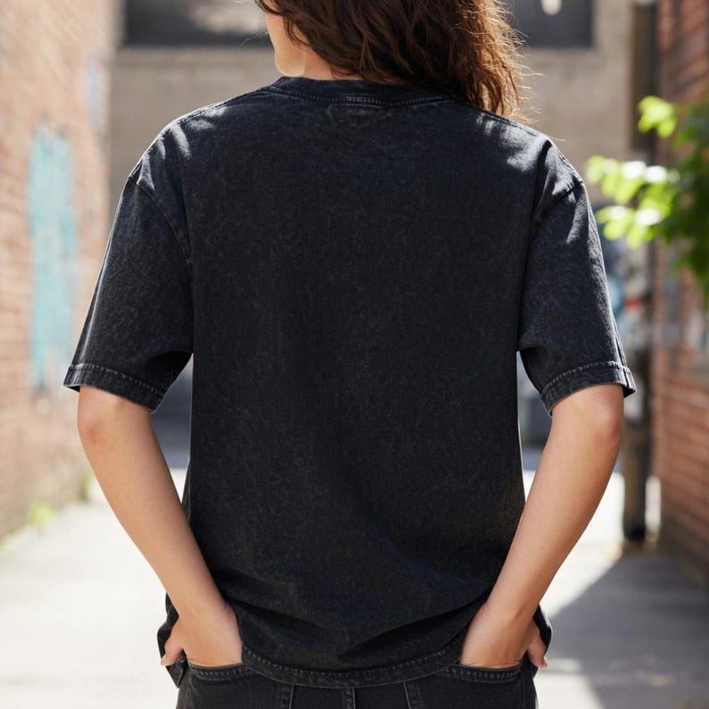 May include: A black, short-sleeved t-shirt with a faded, textured finish. The crew neck shirt is worn by a person with their hands in their pockets. The background shows a brick wall and a narrow alleyway.