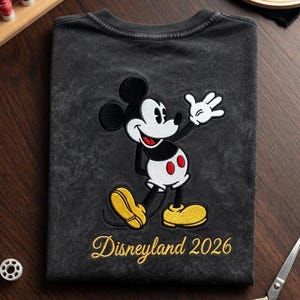 May include: Dark gray t-shirt featuring an embroidered Mickey Mouse design. Mickey is depicted in black, white, red, and yellow. The text "Disneyland 2026" is embroidered in gold. The shirt is on a wooden surface.