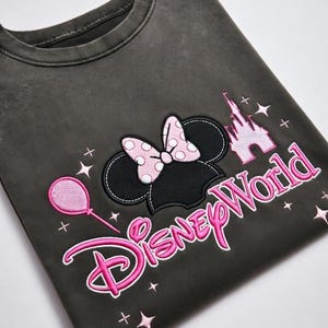 May include: Dark gray sweatshirt featuring a Minnie Mouse design. The design includes Minnie's ears with a pink polka dot bow, a pink castle, a pink balloon, and the words "Disney World" in pink.