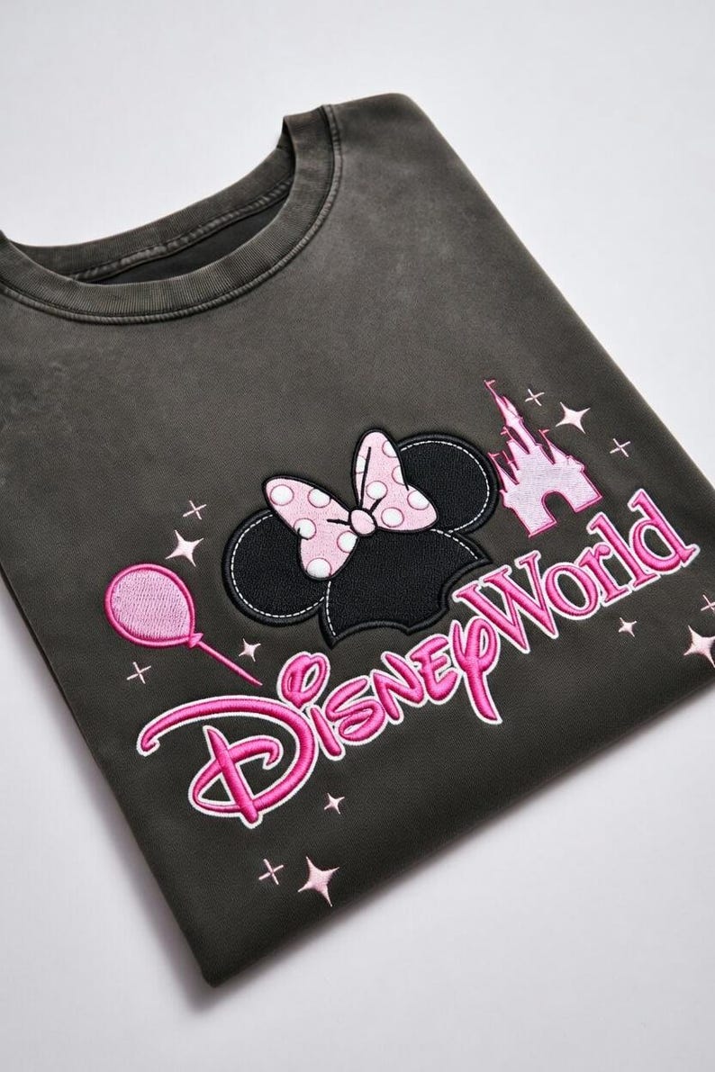 May include: Dark gray sweatshirt featuring a Minnie Mouse design. The design includes Minnie's ears with a pink polka dot bow, a pink castle, a pink balloon, and the words "Disney World" in pink.