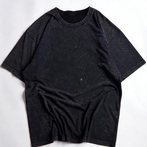 May include: Black short-sleeved t-shirt with a crew neck. The shirt has a distressed, textured appearance with light-coloured streaks throughout. The fabric is draped, and the shirt is laid flat on a white surface.