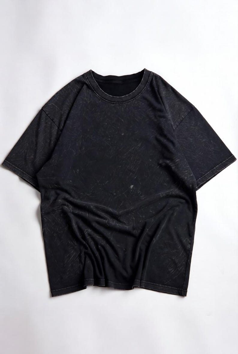 May include: Black short-sleeved t-shirt with a crew neck. The shirt has a distressed, textured appearance with light-coloured streaks throughout. The fabric is draped, and the shirt is laid flat on a white surface.
