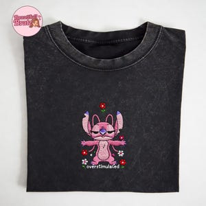 May include: A dark grey t-shirt with an embroidered pink cartoon character with purple details, surrounded by red and white flowers. The text "overstimulated" is below the character. The shirt has a washed-out appearance.
