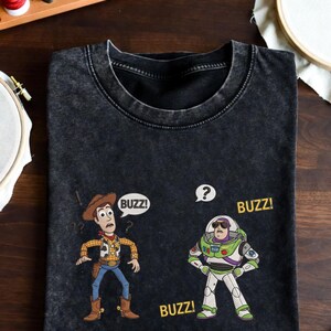 May include: Black t-shirt featuring cartoon characters Woody and Buzz Lightyear from Toy Story. Woody is in a cowboy outfit, and Buzz is in a spacesuit. The word "BUZZ!" appears in speech bubbles and below the characters.