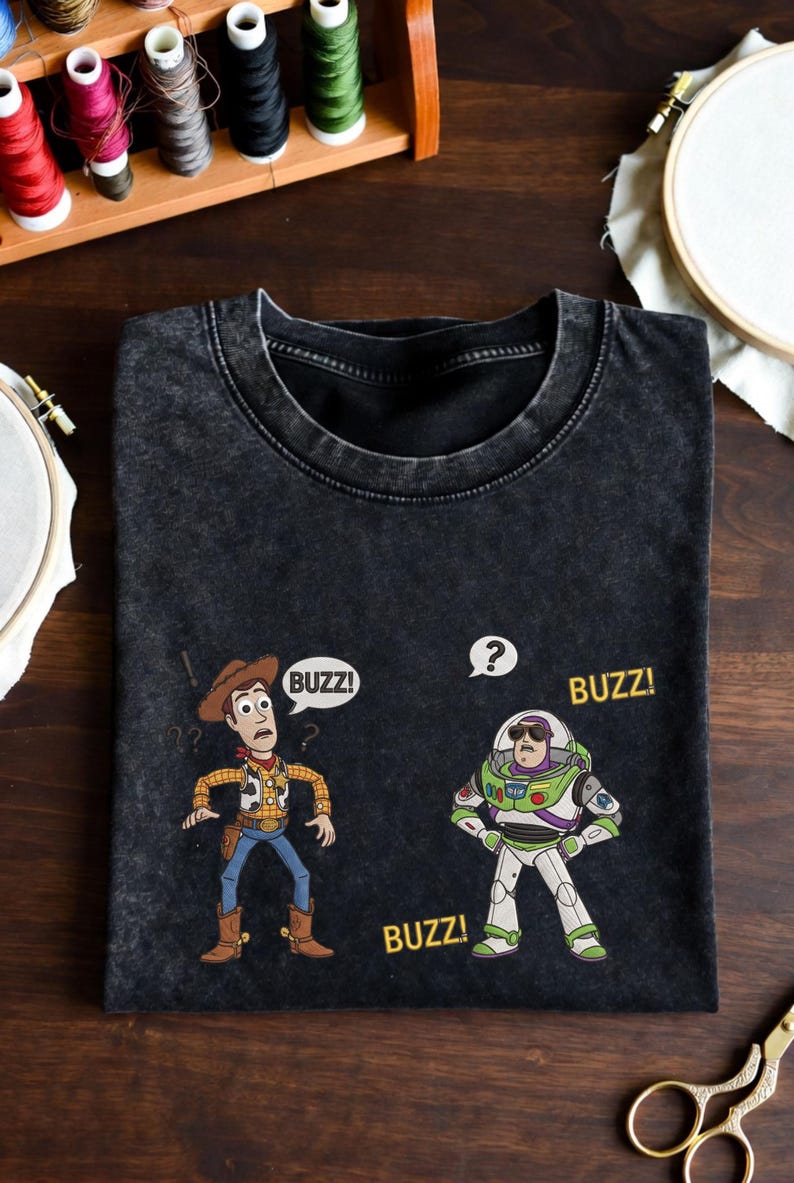 May include: Black t-shirt featuring cartoon characters Woody and Buzz Lightyear from Toy Story. Woody is in a cowboy outfit, and Buzz is in a spacesuit. The word "BUZZ!" appears in speech bubbles and below the characters.