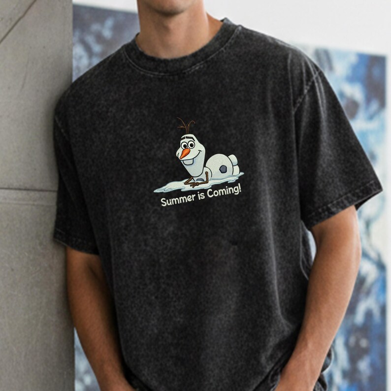 Puede incluir: Camiseta gris oscuro con un dibujo de Olaf, el personaje de dibujos animados, tumbado sobre hielo. El texto "Summer is Coming!" est&aacute; debajo. La camiseta tiene un aspecto vintage descolorido.