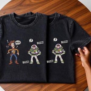 May include: Two black t-shirts with cartoon graphics of Woody and Buzz Lightyear from Toy Story. Woody has question marks, and Buzz has speech bubbles with "BUZZ!" printed on them. The shirts are on a wooden surface.