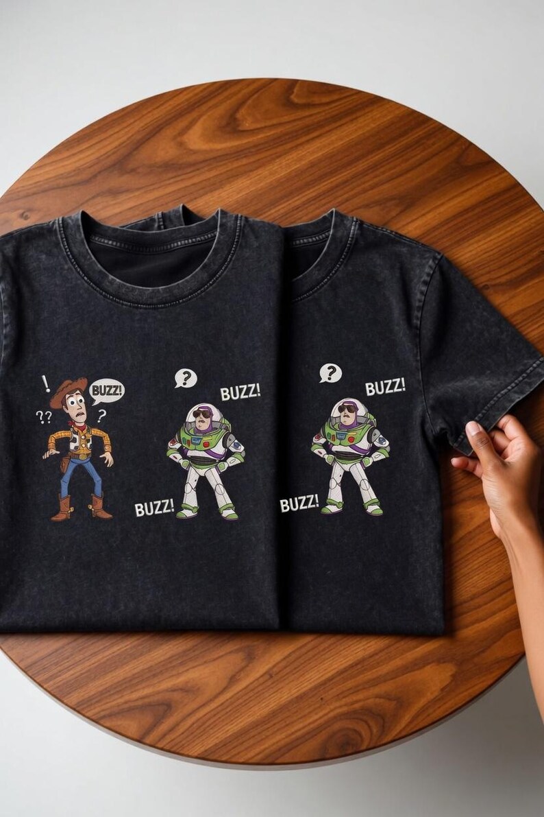 May include: Two black t-shirts with cartoon graphics of Woody and Buzz Lightyear from Toy Story. Woody has question marks, and Buzz has speech bubbles with "BUZZ!" printed on them. The shirts are on a wooden surface.