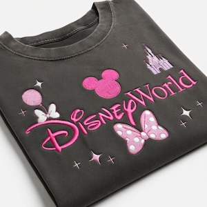May include: Dark gray sweatshirt with pink embroidered "Disney World" text, Mickey Mouse head, castle, bow, and stars. The design includes a balloon and bow. A casual top for Disney fans.