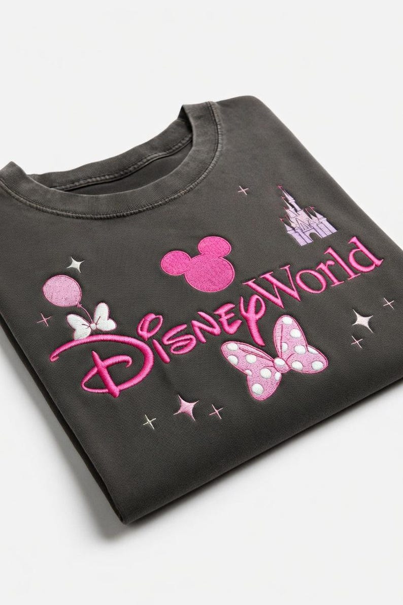 May include: Dark gray sweatshirt with pink embroidered "Disney World" text, Mickey Mouse head, castle, bow, and stars. The design includes a balloon and bow. A casual top for Disney fans.