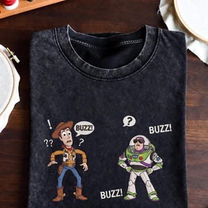 May include: Black t-shirt featuring cartoon characters Woody and Buzz Lightyear. Woody is wearing a cowboy hat and Buzz is in a spacesuit. Speech bubbles say "Buzz!" and "??". The shirt is on a wooden surface with sewing supplies.