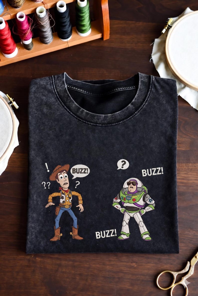 May include: Black t-shirt featuring cartoon characters Woody and Buzz Lightyear. Woody is wearing a cowboy hat and Buzz is in a spacesuit. Speech bubbles say "Buzz!" and "??". The shirt is on a wooden surface with sewing supplies.