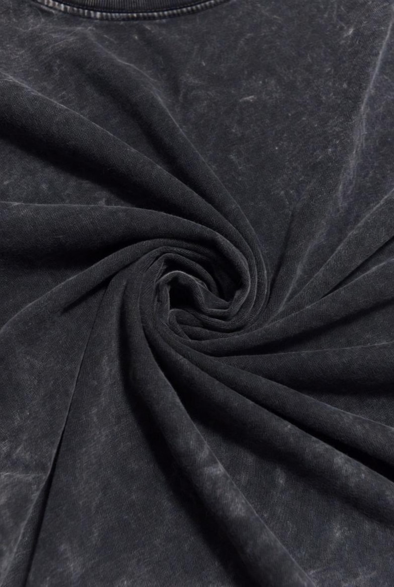 May include: Close-up of a dark grey, acid-washed fabric, possibly a t-shirt. The material is soft and has a textured appearance with lighter areas, creating a vintage or distressed look. The fabric is tightly swirled, showing the texture and drape.