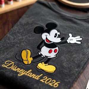 May include: Dark gray t-shirt featuring an embroidered Mickey Mouse design. The classic cartoon character is depicted in black, white, red, and yellow. The words "Disneyland 2026" are embroidered in gold script below the character.