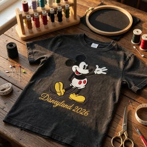 May include: A dark gray t-shirt featuring a Mickey Mouse graphic with yellow shoes and the text "Disneyland 2026" in gold. The image also shows sewing supplies, including spools of thread, scissors, and an embroidery hoop, on a wooden table.