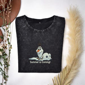 Puede incluir: Camiseta gris oscuro con un dibujo animado de Olaf de Frozen tumbado en un charco de agua. El texto "Summer is Coming!" est&aacute; impreso debajo. Cuello redondo y mangas cortas.