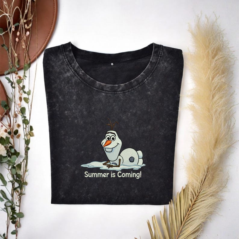 Puede incluir: Camiseta gris oscuro con un dibujo animado de Olaf de Frozen tumbado en un charco de agua. El texto "Summer is Coming!" est&aacute; impreso debajo. Cuello redondo y mangas cortas.
