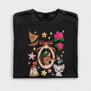 Pode incluir: Camiseta preta com um design bordado com um retrato de uma mulher, um barco, flores, uma estrela do mar e outros elementos tropicais. O design inclui detalhes em rosa, vermelho e amarelo.