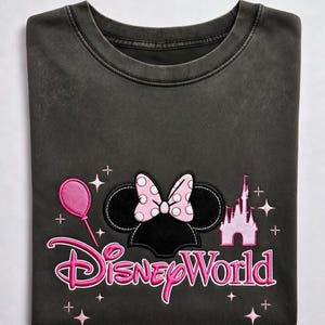 May include: Dark gray t-shirt featuring a Minnie Mouse design with a pink polka-dot bow, a pink balloon, and a pink castle. The words "Disney World" are written in pink script. The shirt is folded.