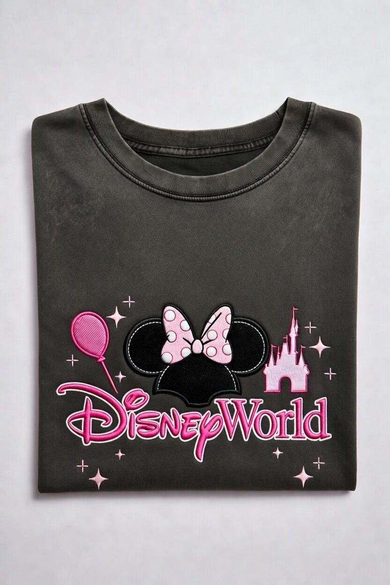 May include: Dark gray t-shirt featuring a Minnie Mouse design with a pink polka-dot bow, a pink balloon, and a pink castle. The words "Disney World" are written in pink script. The shirt is folded.