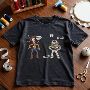 May include: Dark gray t-shirt featuring cartoon characters Woody and Buzz Lightyear from Toy Story. Woody has a speech bubble with "Buzz?" and Buzz Lightyear has a speech bubble with "Buzz!". Sewing supplies, including thread spools, are in the background.