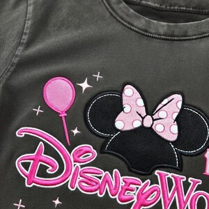May include: Dark gray t-shirt featuring a Minnie Mouse design with a pink polka-dot bow and the words "Disney World" in pink. A pink balloon and stars are also embroidered on the shirt.