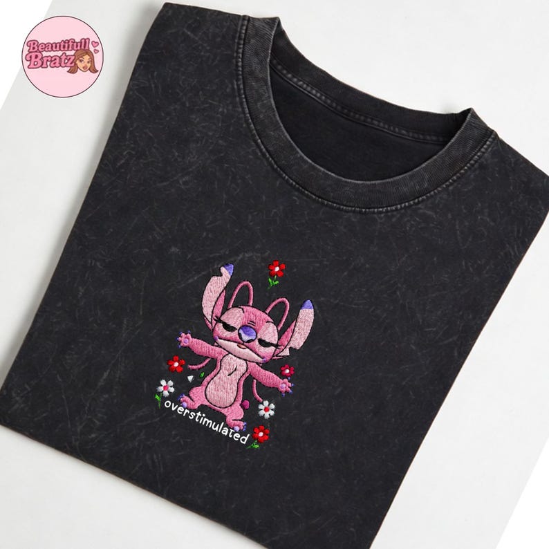 May include: Dark grey t-shirt with an embroidered pink alien character, purple ears, and red and white flowers. The text "overstimulated" is below the character. The shirt has a faded, textured look.