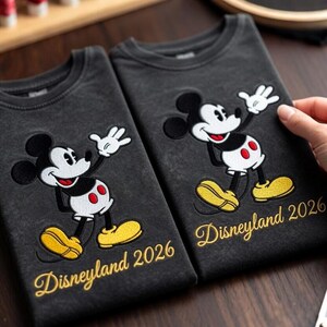 May include: Two dark gray t-shirts with an embroidered Mickey Mouse design. The design features Mickey Mouse in his classic pose with yellow shoes and red shorts. The text "Disneyland 2026" is embroidered in gold.