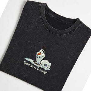 Puede incluir: Camiseta gris oscuro con un dibujo animado de Olaf, tumbado sobre un trozo de nieve. El texto "Summer is Coming!" est&aacute; debajo. La camiseta tiene un lavado vintage.