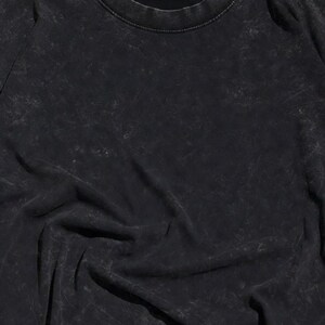 May include: Black crew neck t-shirt with a textured, distressed appearance. The fabric has a faded, worn look, with subtle variations in tone. The shirt's design is simple, with a classic round neckline and long sleeves.