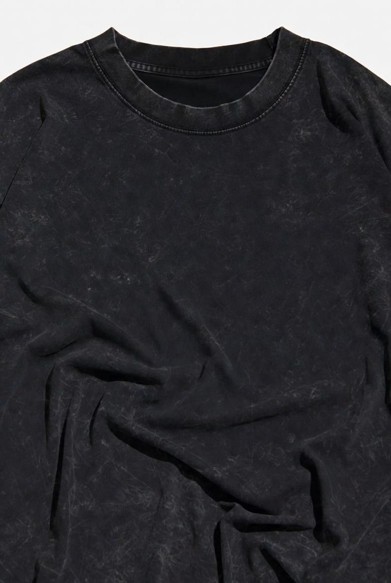 May include: Black crew neck t-shirt with a textured, distressed appearance. The fabric has a faded, worn look, with subtle variations in tone. The shirt's design is simple, with a classic round neckline and long sleeves.