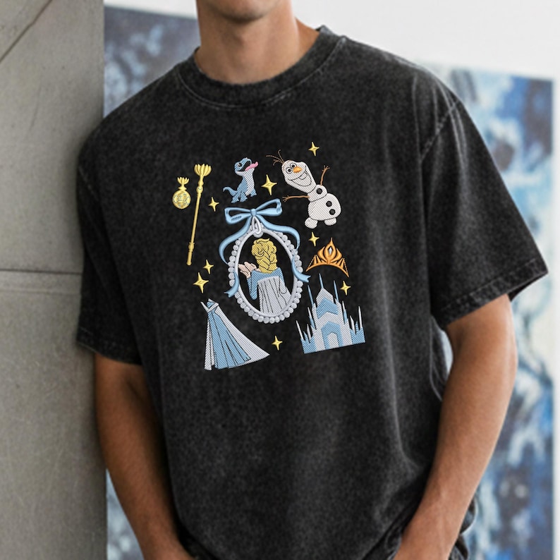 May include: Black t-shirt with embroidered images from the film "Frozen." The design includes Elsa, Olaf, a castle, and other iconic symbols in shades of blue, gold, and white. A fun and colourful design for fans of the animated movie.