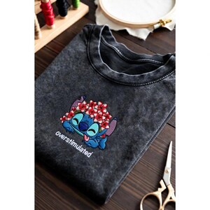May include: Dark gray t-shirt featuring an embroidered Stitch character wearing a flower crown, with the word "overstimulated" below. The design is in shades of blue, red, and white. The shirt is folded on a wooden surface.
