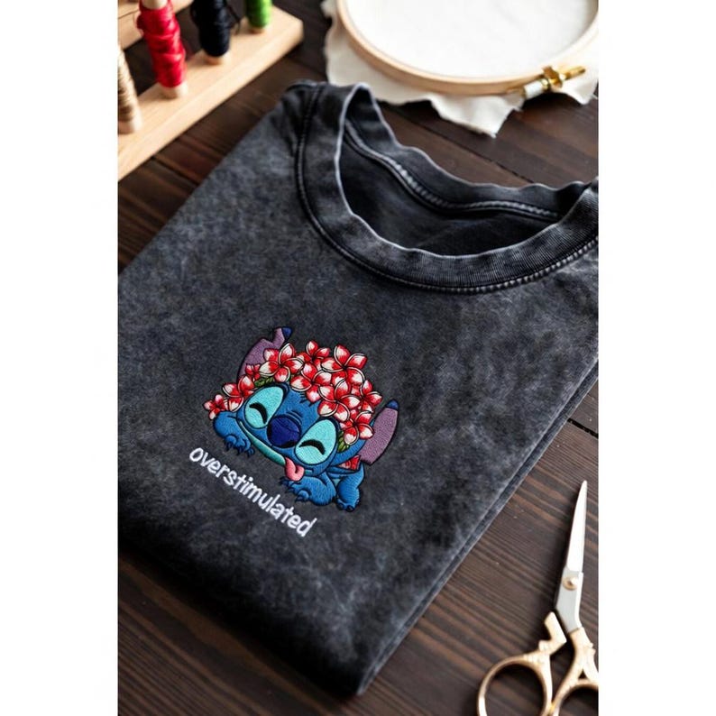 May include: Dark gray t-shirt featuring an embroidered Stitch character wearing a flower crown, with the word "overstimulated" below. The design is in shades of blue, red, and white. The shirt is folded on a wooden surface.