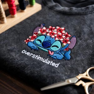 May include: Dark gray t-shirt featuring an embroidered blue cartoon character with a flower crown and the word "overstimulated". The character has a blue body, purple ears, and a pink tongue. The flowers are red and white.