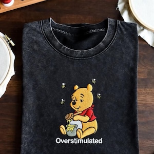 May include: Black t-shirt with an embroidered image of a yellow bear wearing a red shirt, sitting and eating honey. The text "Overstimulated" is below the bear. Sewing supplies and embroidery hoops are in the background.